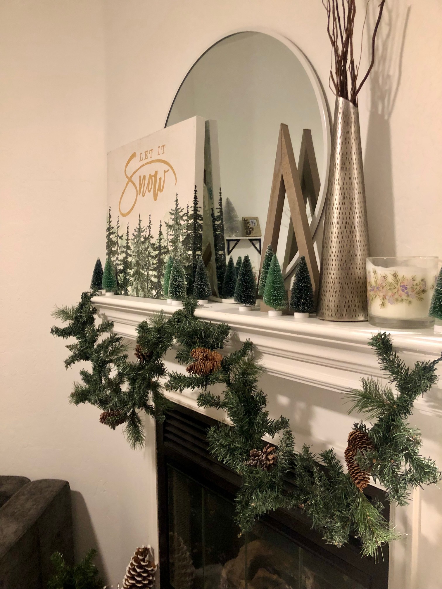 A mantle with various winter decor items such as pine garlands, mini trees, and a large picture of trees in snow. 