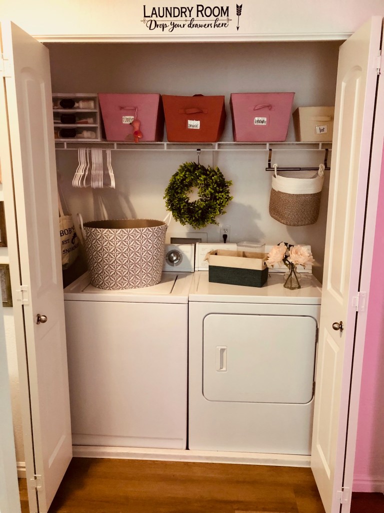 A laundry room done in whites, pinks, and blues with functional baskets and bins with a floral wreath in the middle. 